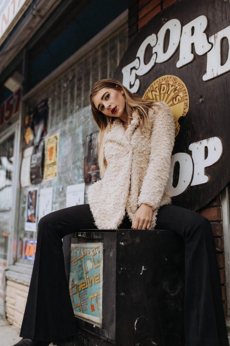 May include: A woman wearing a cream-colored, fuzzy coat and black pants sits on a black box. The background includes a sign that says "RECORD SHOP" and posters on a wall. The overall aesthetic is vintage and stylish.