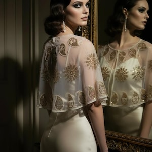Könnte beinhalten: Ein cremefarbenes Satin-Trägerkleid kombiniert mit einem transparenten, kurzärmeligen Capelet. Das Capelet ist mit aufwendigen, goldenen Paillettenstickereien in Blumen- und Paisley-Mustern verziert. Dazu werden lange Perlenohrringe getragen.