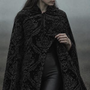 May include: A black velvet cape featuring a tonal damask pattern with a high collar and a tie closure at the neck. The garment is worn over smooth black trousers. The fabric has a textured, raised floral design that contrasts with the dark base.
