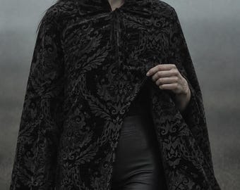 May include: A black velvet cape featuring a tonal damask pattern with a high collar and a tie closure at the neck. The garment is worn over smooth black trousers. The fabric has a textured, raised floral design that contrasts with the dark base.