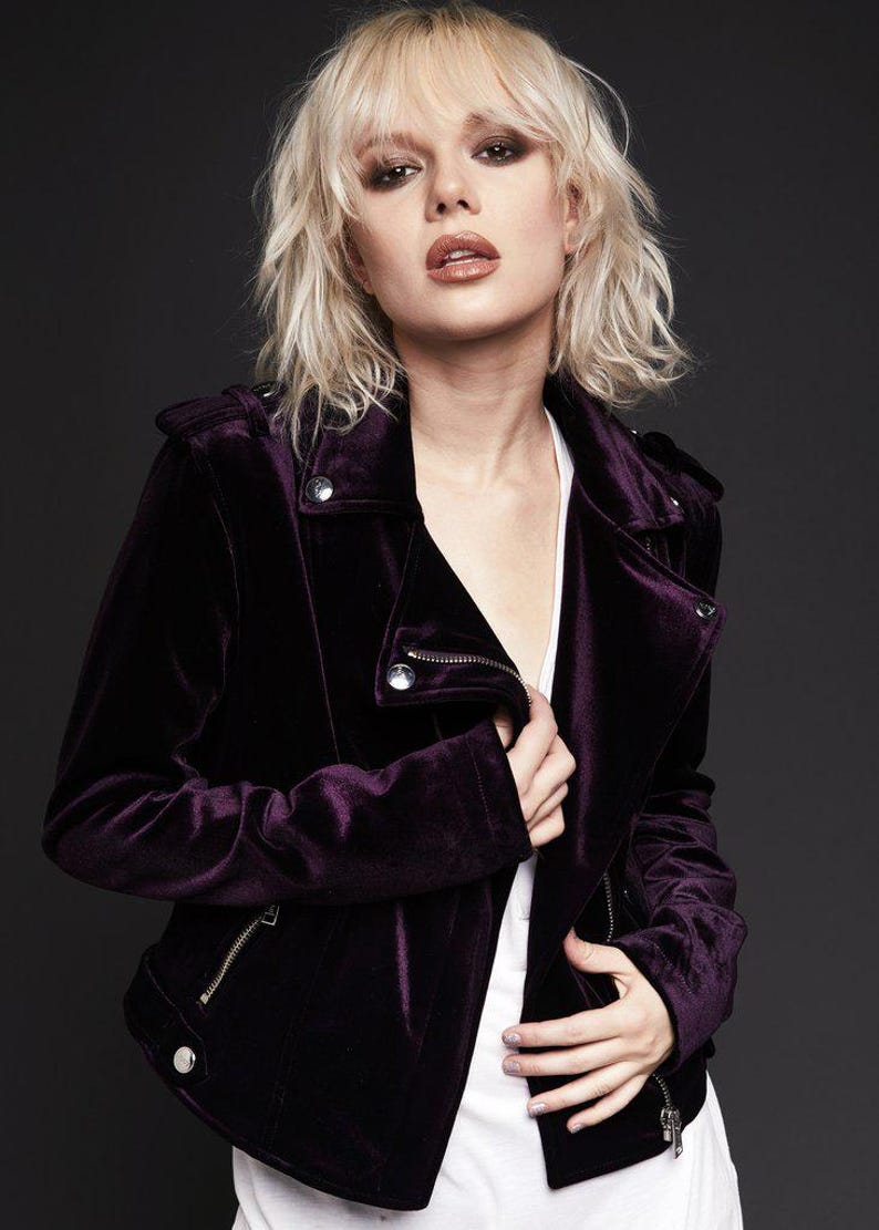 May include: A dark purple velvet jacket with a zipper closure and silver hardware. The jacket has a classic biker style with a notched collar and shoulder epaulets. The model is wearing a white top underneath.