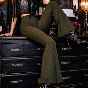 May include: Olive green flared pants paired with black studded high-heeled boots. The pants have a high waist and a wide leg. The boots feature a pointed toe and silver studs. The outfit is styled with a black lace top.