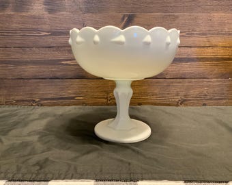 May include: A white milk glass pedestal bowl with a scalloped rim and a raised design of teardrop shapes. The bowl is on a green fabric surface.