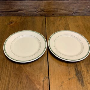 May include: Two white ceramic plates with a green rim. The plates are on a wooden surface.