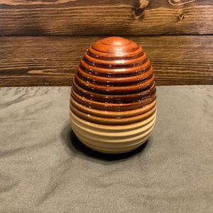 May include: A ceramic egg-shaped container with a brown and white striped pattern. The container is made of glazed ceramic and has a smooth, rounded surface.