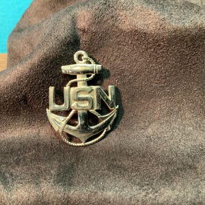 May include: A gold-toned metal pendant featuring a nautical anchor design with the letters USN embossed across the center. A rope detail wraps around the anchor, and a small circular loop is positioned at the top for attachment to a chain.