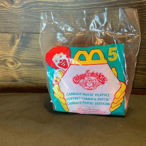 May include: Sealed Cabbage Patch Kids playset in a clear plastic bag. The packaging features a smiling clown, the Cabbage Patch Kids logo, and the number 5. The background is teal and pink with yellow accents.