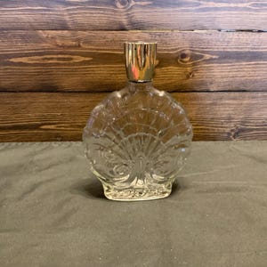 May include: A clear glass bottle with a gold top shaped like a seashell. The bottle has a textured design that resembles a peacock's tail feathers.