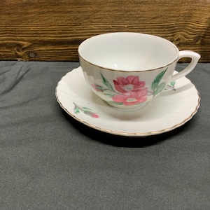 May include: A white teacup and saucer set with a floral design. The teacup and saucer have a gold trim and a pink flower pattern with green leaves. The teacup is on a white saucer with a scalloped edge.