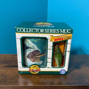 May include: A Rapala Collector Series mug in a green and white box. The box features the text Collector Series Mug and includes a bonus Rapala lure. The mug displays a graphic of a large fish with an open mouth, and the box has a clear window.