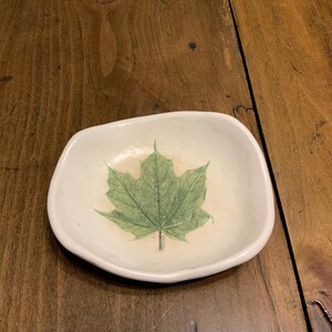 May include: A white ceramic dish with a green leaf design. The dish is slightly oval shaped and has a raised rim.