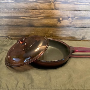 May include: A red glass skillet with a matching lid. The skillet has a long handle and a smaller handle on the opposite side. The lid has a knob on top.