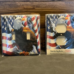 May include: Two decorative switch plates featuring an American bald eagle with an American flag background. The switch plates are made of plastic and have a glossy finish. The text on the bottom of the switch plate reads "Switchplate Gallery Matching Hand-Painted Screws Included!"