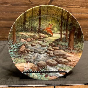 May include: A ceramic plate with a woodland scene featuring a stream, rocks, and two deer. The plate is decorated with green, brown, and white colors. The plate is signed "JERNER" in the lower right corner.