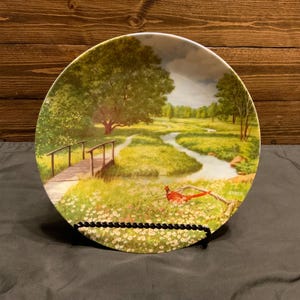 May include: A white ceramic plate with a painted scene of a winding stream through a green meadow with a wooden bridge and a red bird. The plate is on a black stand.