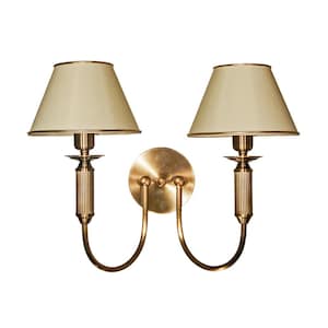 May include: A double wall sconce with a brass finish. It has two cream-coloured lampshades with gold trim. The fixture features a decorative design with curved arms and a round backplate, suitable for wall mounting.