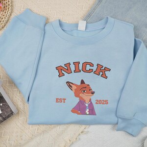 May include: A pale blue sweatshirt with the name "NICK" in orange block letters and a fox illustration. The text "EST 2025" is below the fox. The sweatshirt is styled with jeans and trainers.
