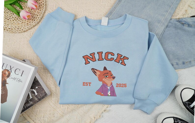 May include: A pale blue sweatshirt with the name "NICK" in orange block letters and a fox illustration. The text "EST 2025" is below the fox. The sweatshirt is styled with jeans and trainers.