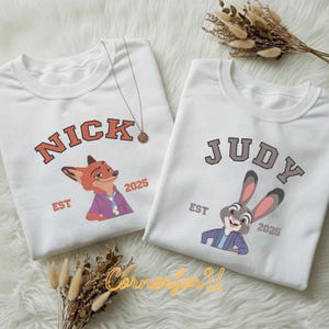 May include: Two white crew neck sweatshirts. One has "NICK" in orange block letters above a cartoon fox, the other says "JUDY" above a cartoon bunny. Both have "EST 2025" below the characters. A gold necklace is visible.