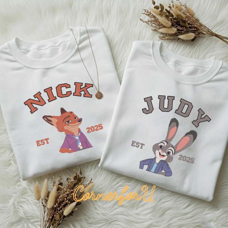 May include: Two white crew neck sweatshirts. One has "NICK" in orange block letters above a cartoon fox, the other says "JUDY" above a cartoon bunny. Both have "EST 2025" below the characters. A gold necklace is visible.