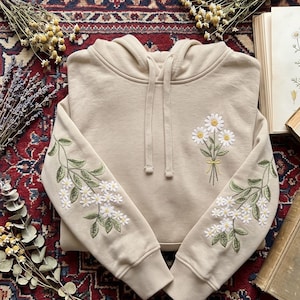 May include: A beige hooded sweatshirt featuring embroidered floral designs. A small bouquet of white daisies with yellow centres is stitched on the chest, while both sleeves display long, trailing stems with green leaves and white daisy blossoms.