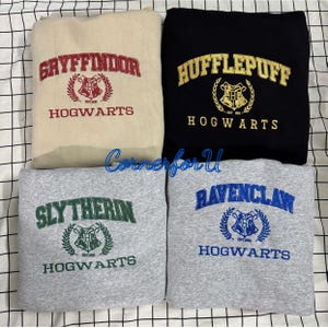 Peut inclure: Quatre sweat-shirts avec des textes et des écussons brodés. Les sweat-shirts sont beiges, noirs et deux nuances de gris. Les textes incluent "Gryffindor", "Hufflepuff", "Slytherin" et "Ravenclaw", avec "Hogwarts" en dessous.