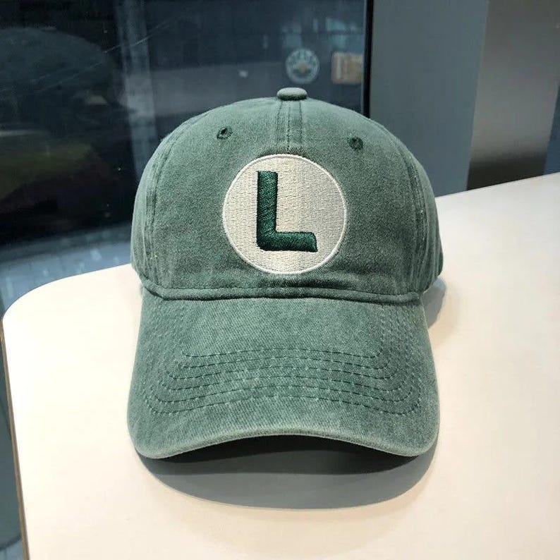May include: A green baseball cap with a curved brim. The cap features a white circular patch with a green embroidered letter "L". The cap is made of a soft, worn material.