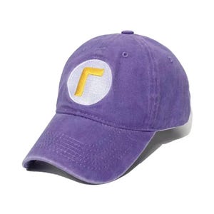 May include: A purple baseball cap with a curved brim. The cap features a white circle with a yellow "L" shape embroidered on the front. The cap is made of a soft, textured fabric.