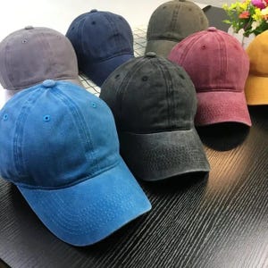 May include: A collection of baseball caps in various colors, including blue, gray, black, and burgundy. The caps have a classic design with a curved brim and adjustable strap. The fabric appears to be a washed cotton.