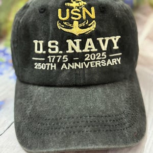 Könnte beinhalten: Schwarze Baseballkappe mit dem Text "USN" und einem goldenen Ankeremblem. Die Kappe trägt auch den Text "U.S. NAVY - 1775 - 2025 250TH ANNIVERSARY" in Weiß. Die Kappe ist aus weichem, abgenutztem Stoff gefertigt.