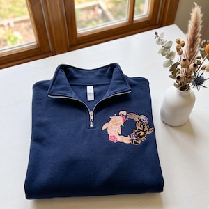 Könnte beinhalten: Ein marineblaues Sweatshirt mit Viertelreißverschluss und einer Stickerei auf der Brust. Das Motiv zeigt zwei Koi-Fische in kreisförmiger Anordnung, einer in Hellrosa und einer in Schwarz, verziert mit kleinen Blumen, Sternen und Mondformen.