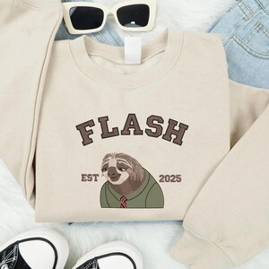 May include: A cream-coloured sweatshirt featuring the word "FLASH" in brown lettering above a cartoon sloth wearing a green shirt and red tie. The text "EST 2025" is below the sloth. Sunglasses and trainers are also in the image.