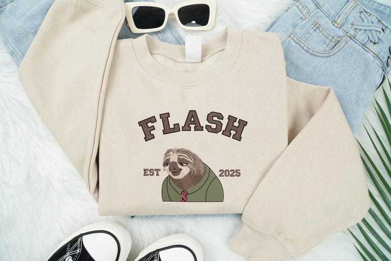 May include: A cream-coloured sweatshirt featuring the word "FLASH" in brown lettering above a cartoon sloth wearing a green shirt and red tie. The text "EST 2025" is below the sloth. Sunglasses and trainers are also in the image.