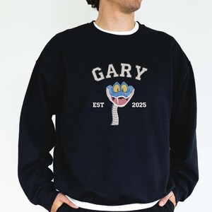 May include: Dark blue sweatshirt with a white crew neck. The front displays the word "GARY" in white arched lettering above a cartoon snake with blue eyes and a pink tongue. Below the snake are the words "EST 2025".