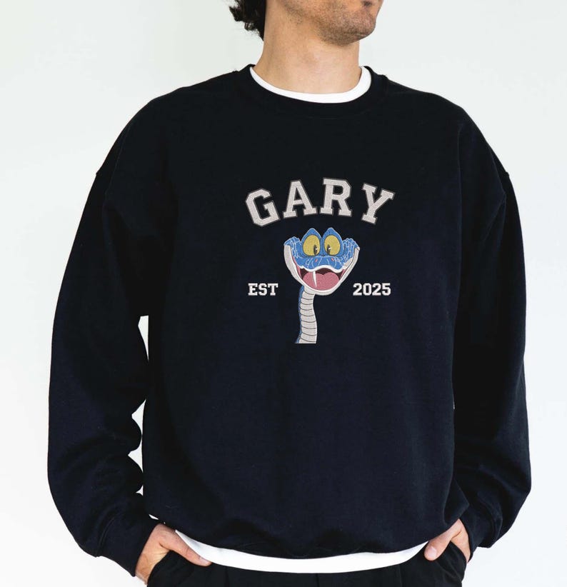 May include: Dark blue sweatshirt with a white crew neck. The front displays the word "GARY" in white arched lettering above a cartoon snake with blue eyes and a pink tongue. Below the snake are the words "EST 2025".
