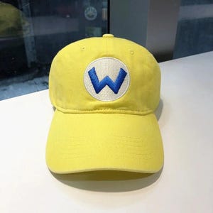 May include: Yellow baseball cap with a curved brim. The cap features a white circular patch with a blue "W" embroidered on the front. The cap is made of a soft material and has a button on top.
