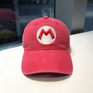 May include: A red baseball cap featuring a white circle with a red "M" logo. The cap has a curved brim and a fabric strap closure. The hat is a casual accessory.