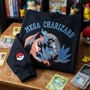 Könnte beinhalten: Ein schwarzer Kapuzenpullover mit einer Stickerei von Mega Glurak und dem Schriftzug "MEGA CHARIZARD" in Hellblau. Am Ärmel befindet sich ein kreisförmiger Aufnäher in Rot, Weiß und Schwarz in Form eines Pokéballs. Das Motiv zeigt orange und blaue Drachen.