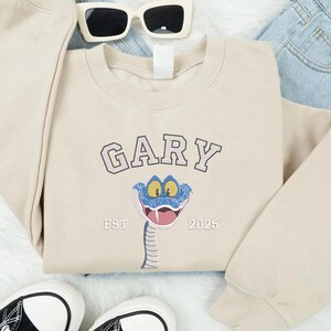 May include: A cream-coloured sweatshirt featuring the name "GARY" in a collegiate font above a cartoon snake with a blue head and a pink tongue. The text "EST 2025" is below the snake. The sweatshirt is styled with sunglasses, jeans, and trainers.
