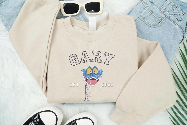 May include: A cream-coloured sweatshirt featuring the name "GARY" in a collegiate font above a cartoon snake with a blue head and a pink tongue. The text "EST 2025" is below the snake. The sweatshirt is styled with sunglasses, jeans, and trainers.