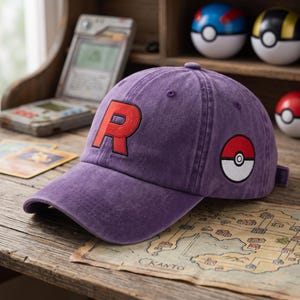 Könnte beinhalten: Eine violette Baseballkappe mit einem großen, aufgestickten roten Buchstaben R auf der Vorderseite und einem kreisförmigen rot-weiß-schwarzen Pokéball-Emblem an der Seite. Die Kappe hat einen gebogenen Schirm und eine strukturierte Krone mit Ösen.