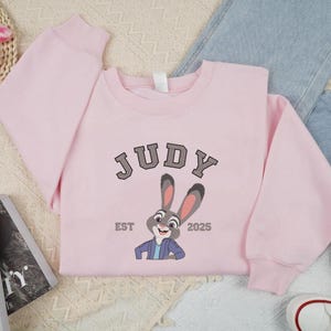 May include: A pale pink sweatshirt with the name "JUDY" in grey block letters and a cartoon rabbit graphic. The text "EST 2025" is below the rabbit. The sweatshirt is laid flat with jeans and trainers.