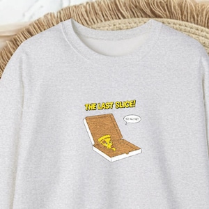 Könnte beinhalten: Hellgraues Sweatshirt mit einer Grafik einer Pizzaschachtel mit dem letzten Pizzastück. Der Text "THE LAST SLICE!" befindet sich über der Pizzaschachtel. Das Sweatshirt liegt auf einer gewebten Matte.