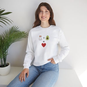 Könnte beinhalten: Weißes Sweatshirt mit einer Grafik einer Katze plus einer Avocado, die ein rotes Herz ergibt. Die Katze ist orange und weiß, und die Avocado ist grün. Das Sweatshirt hat lange Ärmel.