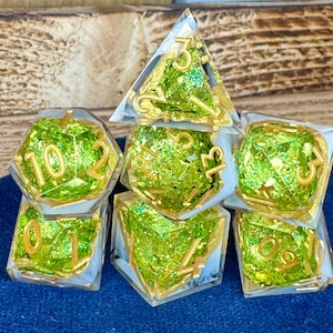 May include: A set of seven polyhedral dice. Each die is clear with green glitter and gold numbering. The dice are arranged on a blue surface. The dice are for tabletop games.