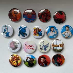 Puede incluir: Una colección de botones redondos con varios diseños. Algunos botones presentan arte abstracto en rojo, naranja y azul. Otros muestran imágenes de personas, personajes de dibujos animados y los textos "SLOW DOWN" y "Wake Up!".