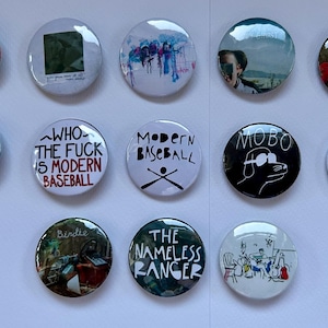 May include: A collection of 12 round button pins with various designs. The pins feature a variety of colors, including black, white, red, blue, and green. Some of the pins have text on them, such as "Who the Fuck is Modern Baseball" and "The Nameless Ranger".