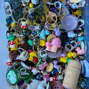 Pode incluir: Uma coleção de bugigangas e pequenos brinquedos coloridos em um recipiente de plástico transparente. Os itens incluem pequenos animais de plástico, botões, tampas de garrafas, dados, anéis e outros itens diversos.
