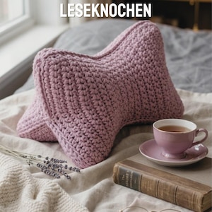 May include: A handmade, light purple crochet pillow with the text "HÄKELANLEITUNG LESEKNOCHEN" and "Hand-Made-Gifts by Iwona". A teacup and saucer, a book, and lavender sprigs are also visible.