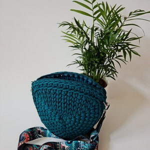 Könnte beinhalten: Tealfarbene runde Umhängetasche aus Häkelarbeit mit einem gemusterten Riemen. Die Tasche liegt auf einer weißen Oberfläche mit einer grünen Pflanze im Hintergrund.